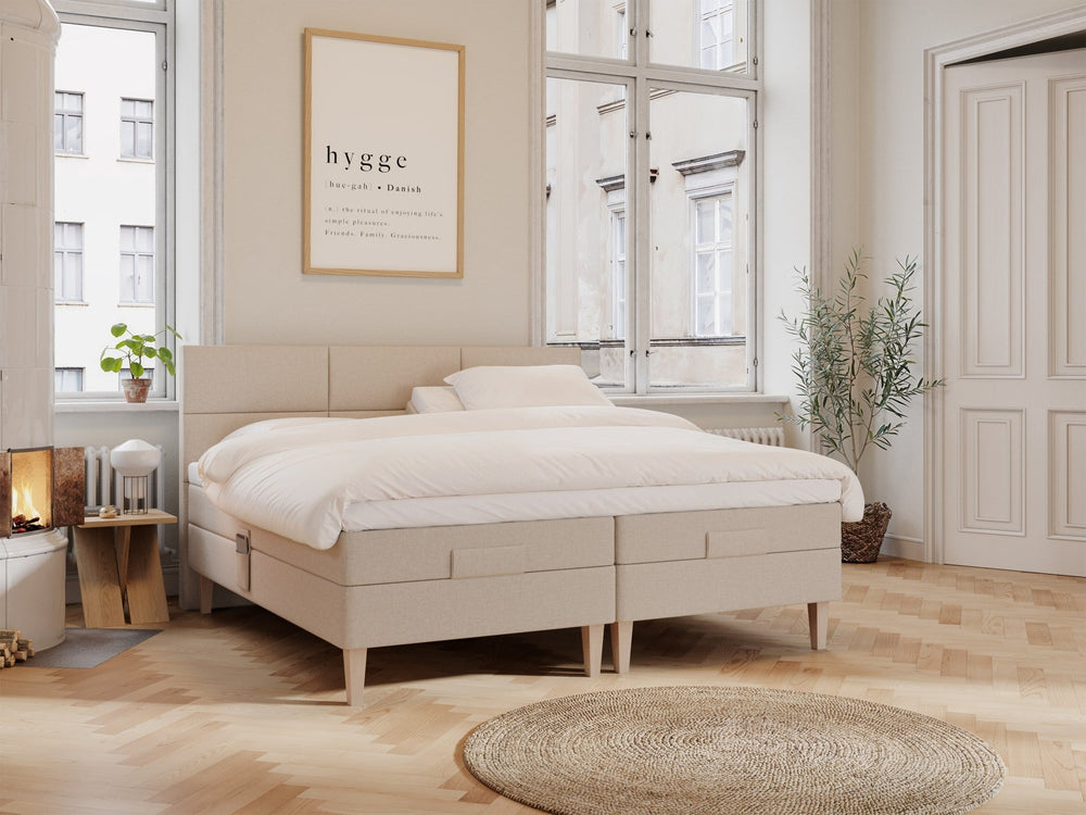 Sandfarget regulerbar dobbeltseng Ragna 210x210 med myk polstret ramme, hvit dyne og puter i lyst, minimalistisk soverom.