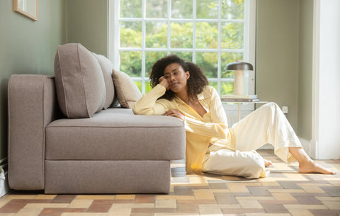 Beige stoffsofa med runde puter, lys pudderpute og person som lener seg mot sofa i gult plagg ved vindu