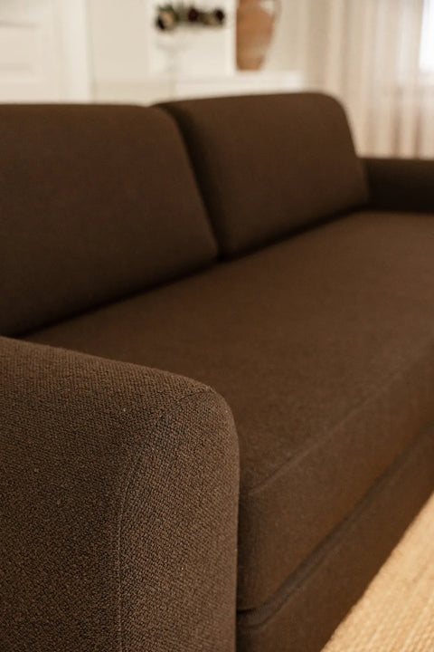 Nærbilde av brun sofa i grovt vevd stoff, fokus på armlene og puterygg i moderne, rektangulær form