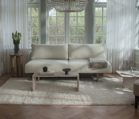 Lys kremfarget sofa med mykt tekstil, plysjteppe, tynn pledd og hvite gardiner foran vindu.