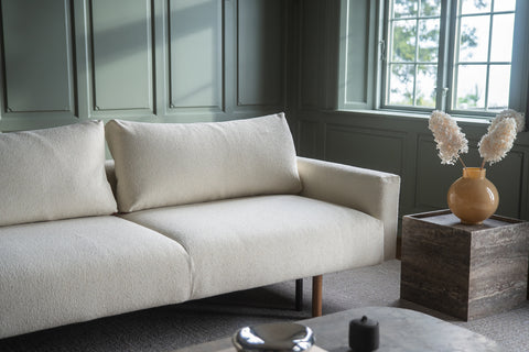Lyse kremfarget sofa i boucle-stoff med firkantede puter, grønn panelvegg og gul keramikkvase med tørkede blomster