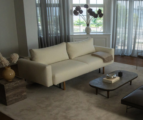 Lys kremfarget sofa i mykt tekstil med matchende pute og beige pledd i stilig stue ved vindu